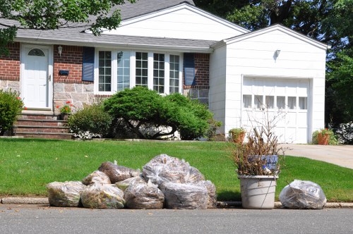 A row of large, irregularly shaped plastic rubbish sacks filled with waste, positioned along the curb in front of a residential driveway in a suburban area. The sacks are translucent with a greyish appearance, and some have visible contents. To the right of the sacks, there is a metal planter containing dead or dried plants, indicating recent outdoor clutter. The background features a brick and white siding house with a bay window, a white garage door, a well-maintained front garden with trimmed bushes and grass, and a paved driveway. The scene is illuminated by natural daylight, suggesting a clear day, and the overall environment appears clean and tidy, consistent with a typical rubbish collection point in a residential suburb near Finsbury Park.