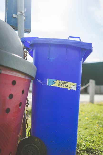 A black wheeled rubbish bin positioned on a residential pavement at night, with the label 'ST. JOHN'S' visible on the front. The bin is partially open and filled with various waste materials, includin